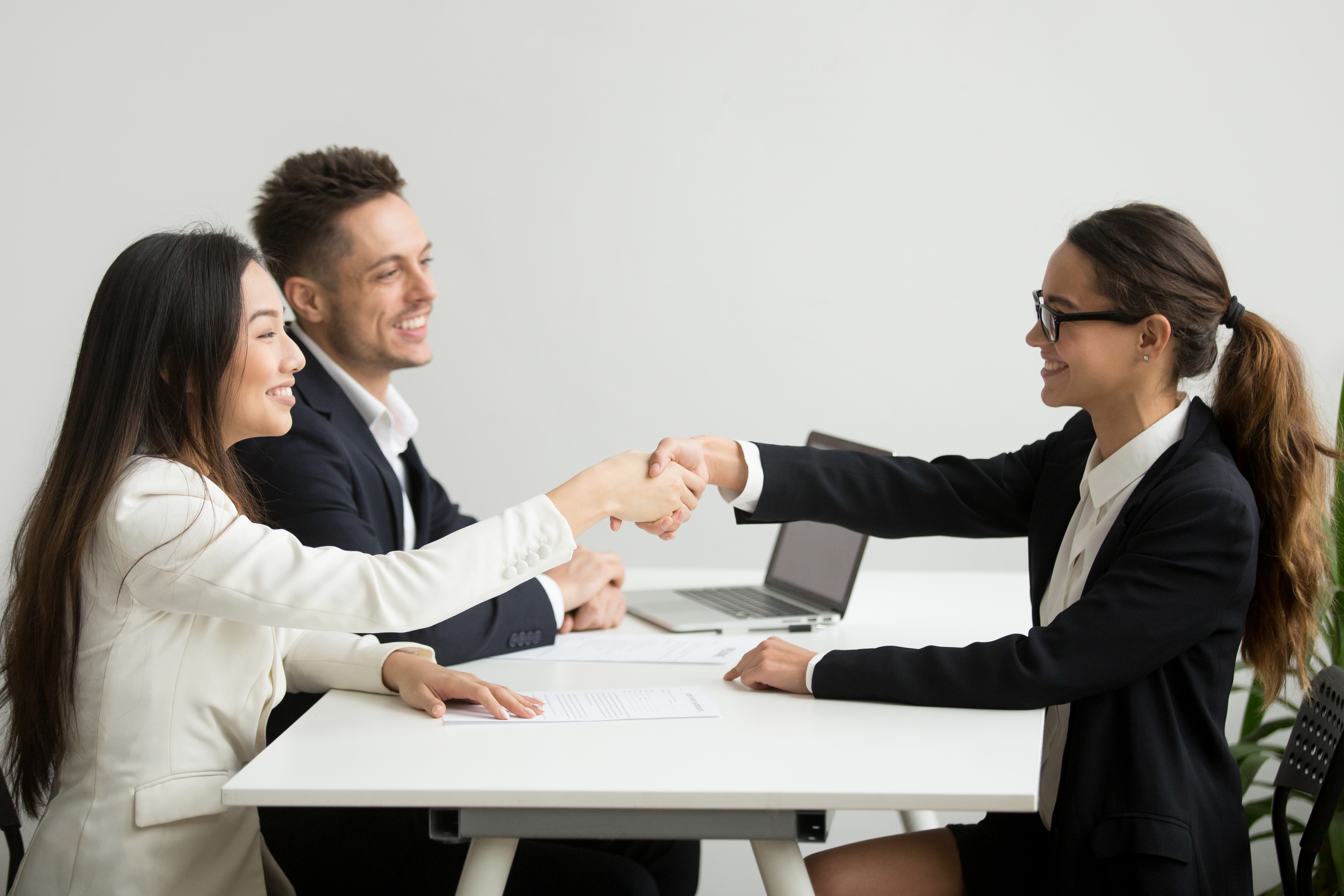 Smiling diverse businesswomen shake hands at group meeting, friendly asian hr handshaking congratulating hired applicant at job interview, satisfied millennial partners make contract deal concept